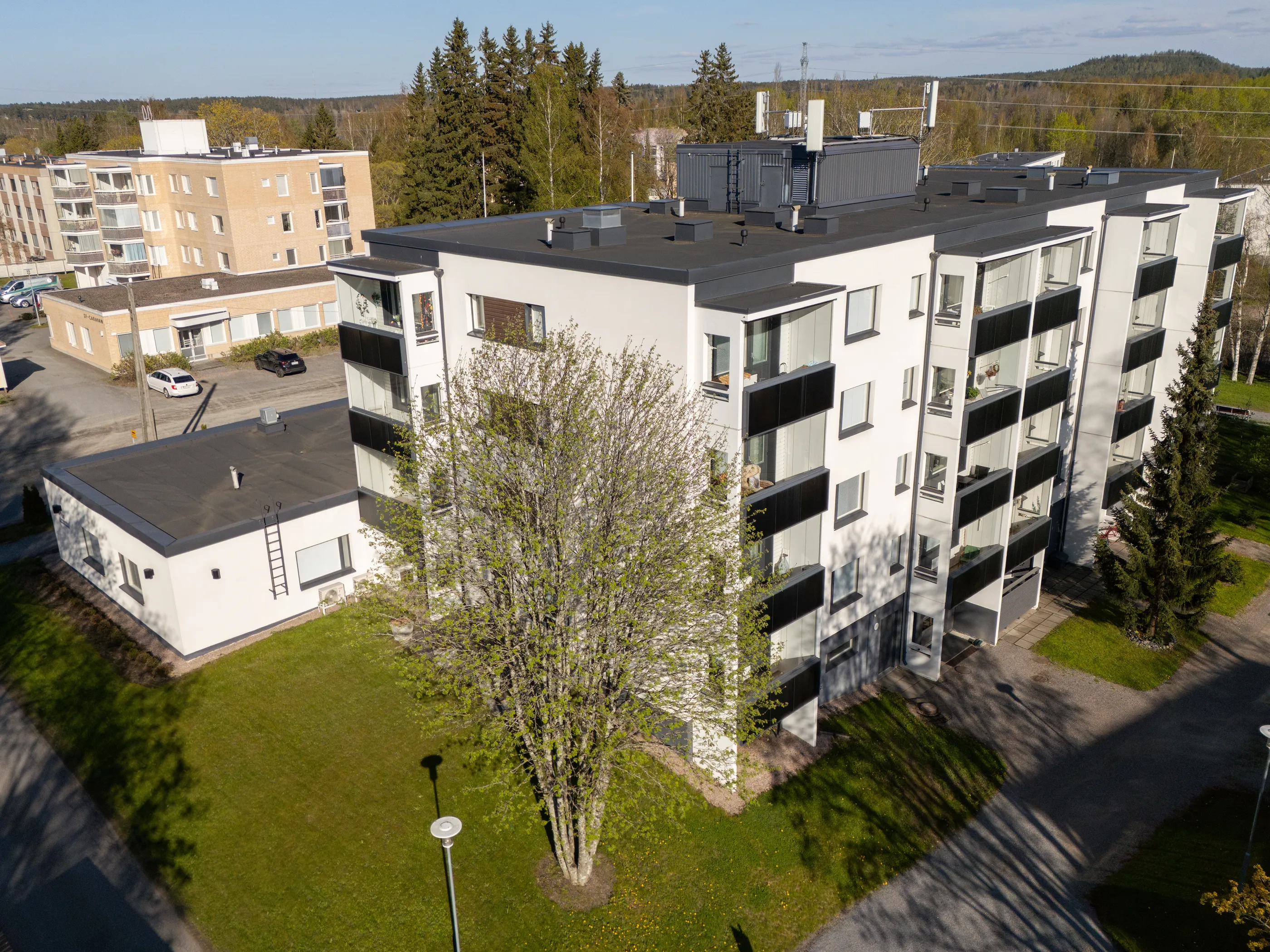 Ilmakuva aurinkoisesta asuinalueesta. Edustalla on moderni, valkoinen kerrostalo tummanharmailla yksityiskohdilla ja lasitetuilla parvekkeilla. Sen edessä kasvaa puu, jossa on uusia vihreitä lehtiä. Taustalla näkyy vaaleanruskea kerrostalo ja pienempi rakennus. Maisemassa on vihreää nurmikkoa, havupuita ja lehtipuita sekä teitä, joilla pitkät varjot korostavat auringonpaistetta. Kaukana horisontissa on metsäinen kukkula ja matkapuhelintorni.