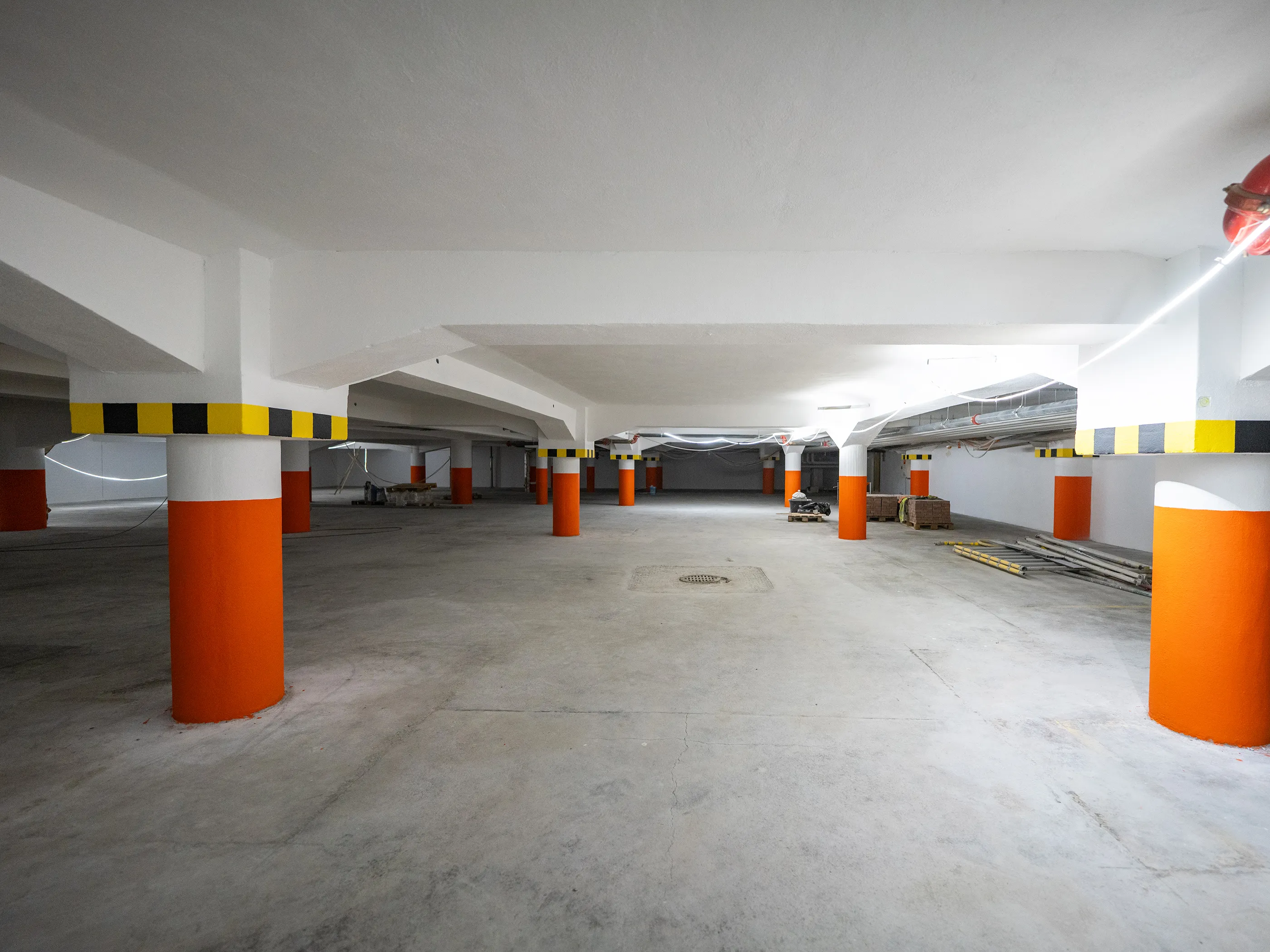 Tilava, kirkkaasti valaistu maanalainen tila, mahdollisesti parkkihalli tai kellari. Useat pyöreät betonipylväät ovat alaosastaan oransseja ja yläosastaan valkoisia. Pylväiden yläpuolella olevissa palkeissa on musta-keltaiset varoitusraidat. Lattia on harmaata betonia ja katto valkoinen, näkyvillä johdotuksilla ja valoilla. Taustalla on rakennustarvikkeita, kuten lavoja, tiiliä ja tikkaita. Etualalla lattiassa näkyy kaivonkansi. Oikeassa yläkulmassa on punainen paloturvallisuuslaite.