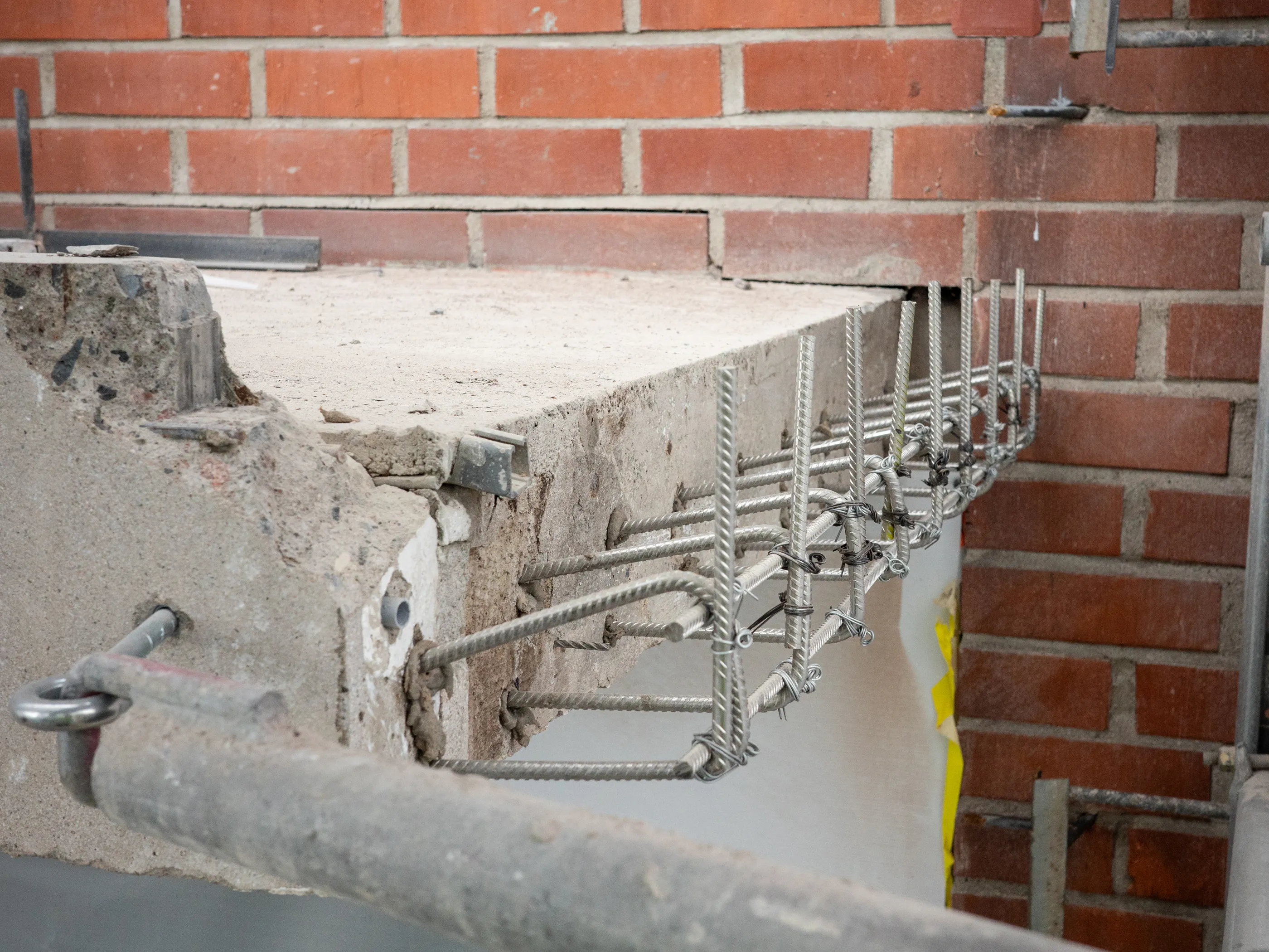 Lähikuva rakennustyömaasta, jossa betonipalkkia tai -laattaa vahvistetaan. Betonin etuosa on rikki, paljastaen sen karkean pinnan ja kiviaineksen. Siitä ulottuu esiin teräksisiä harjateräksiä, jotka on sidottu yhteen ohuella rautalangalla muodostaen raudoituskehikkoja. Taustalla näkyy punatiiliseinä. Betonissa on myös upotettuna erilaisia metalliosia, kuten putki ja silmukkapultti. Kuva on hyvin valaistu, korostaen materiaalien yksityiskohtia.