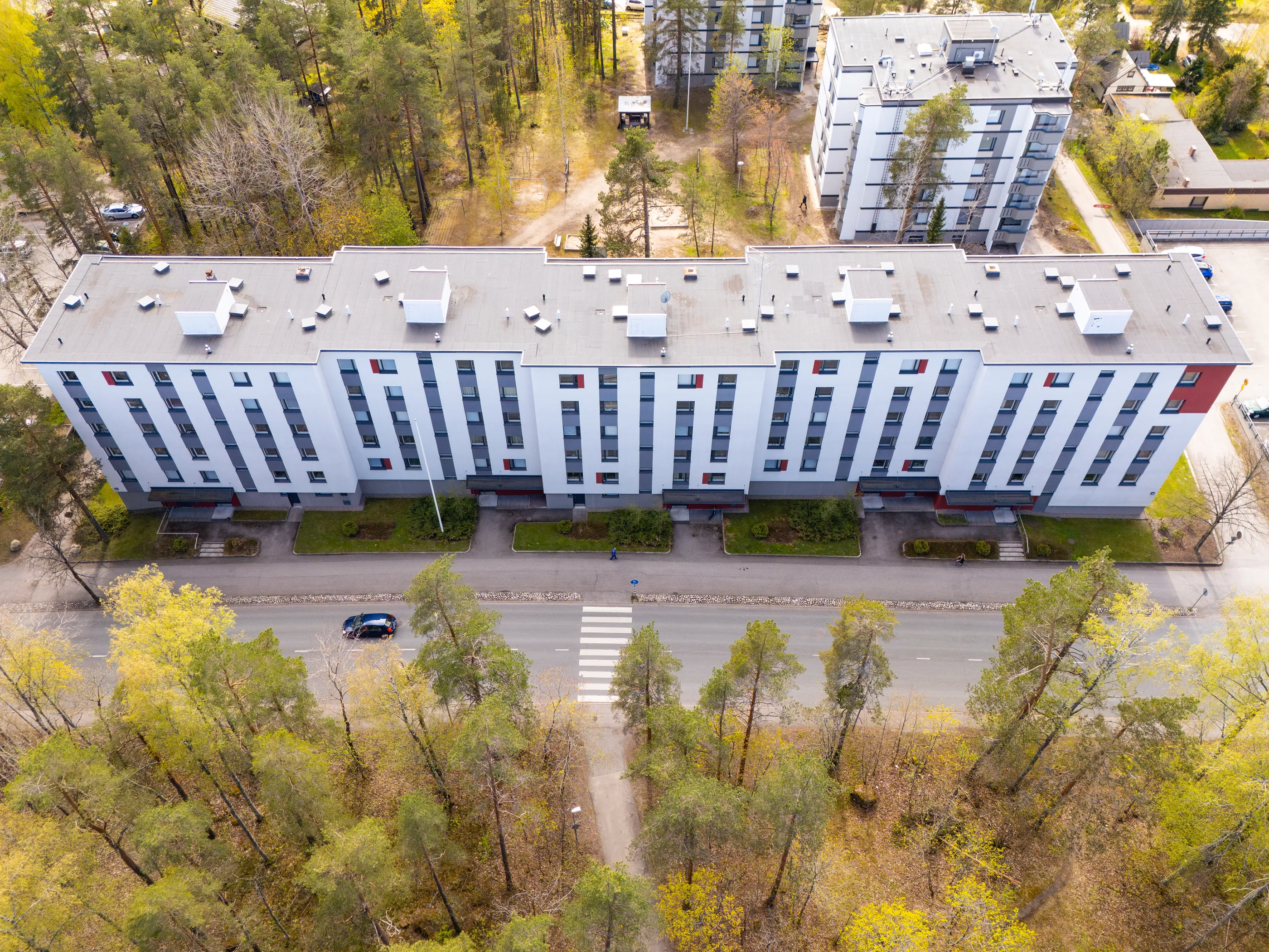 Ilmakuva asuinalueesta, jossa on pitkä, valkoinen kerrostalo harmailla ja punaisilla yksityiskohdilla. Talon edessä kulkee tie suojatien ja tummansinisen auton kanssa. Tien toisella puolella ja rakennuksen takana on metsäinen alue, jossa on havupuita ja kevään vihreitä lehtipuita. Muutama ihminen kävelee poluilla ja jalkakäytävillä. Taustalla näkyy muita kerrostaloja ja pientaloja. Kuva on otettu kirkkaassa päivänvalossa.