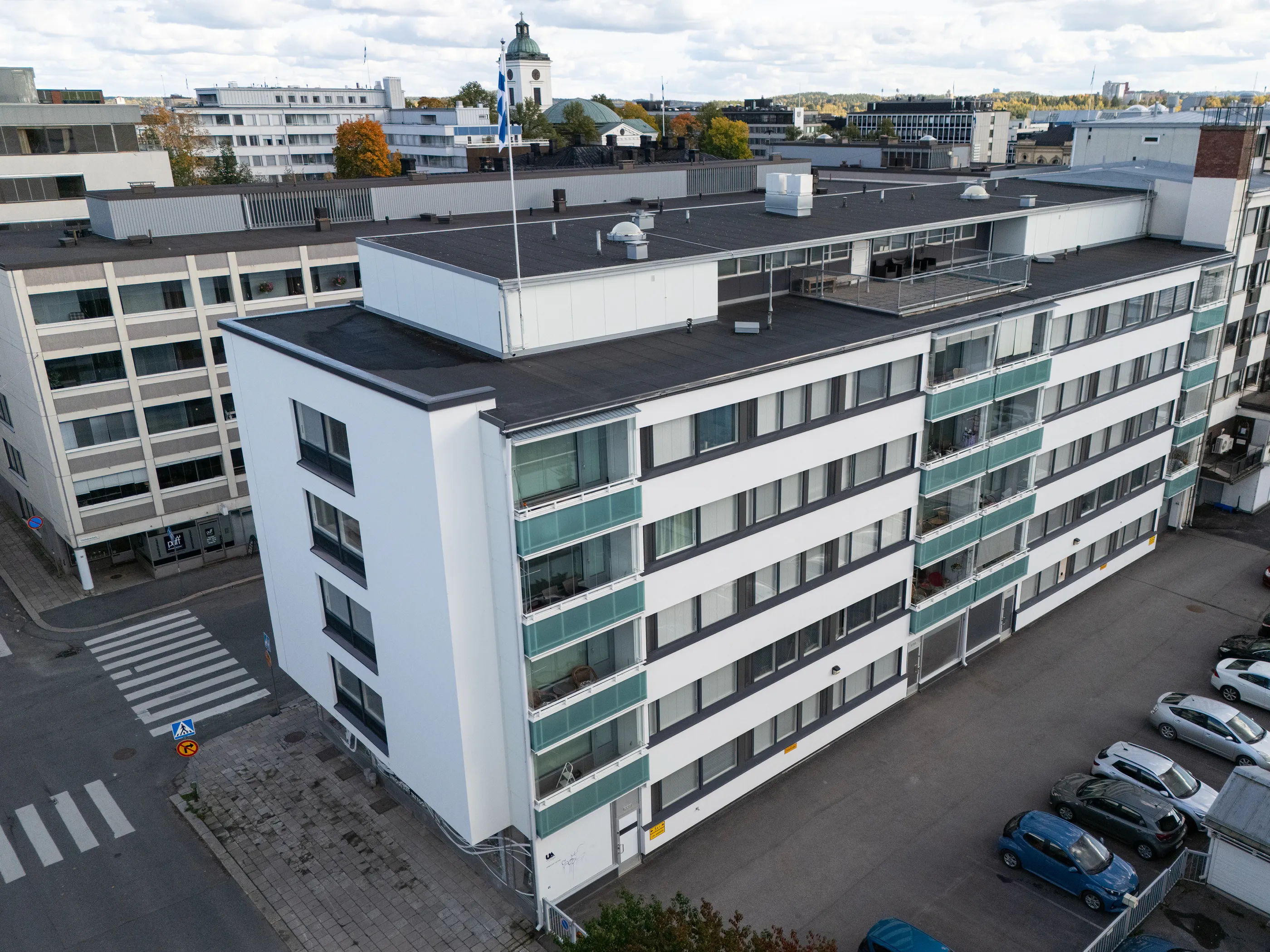 Yläkuva modernista valkoisesta asuinkerrostalosta, jossa on tummat ikkunat ja vihreäsävyiset lasiparvekkeet. Rakennuksen edessä on parkkipaikka useine autoineen ja vasemmalla katu suojateineen. Taustalla näkyy kaupunkimaisema, jossa on muita rakennuksia, syksyisiä puita ja kirkontorni Suomen lipun kanssa. Taivas on pilvinen.