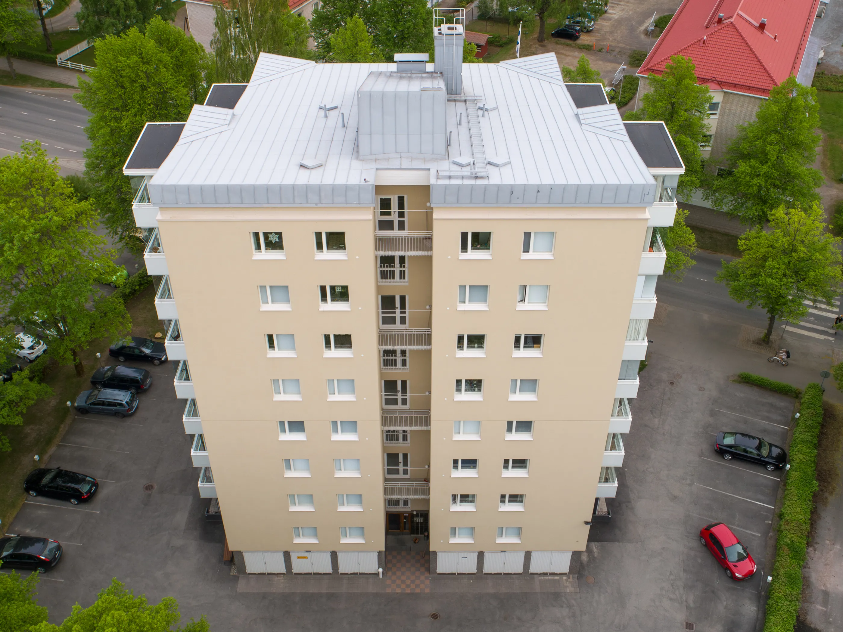 Ylhäältä otettu kuva vaaleanbeigestä kerrostalosta, jossa on hopeanvärinen peltikatto ja valkoiset parvekkeet. Rakennuksen ympärillä on vihreitä puita, pensaita ja pysäköintialueita, joilla on useita autoja. Taustalla näkyy tie suojateineen ja punatiilikattoinen rakennus. Kuva on otettu kirkkaassa päivänvalossa.