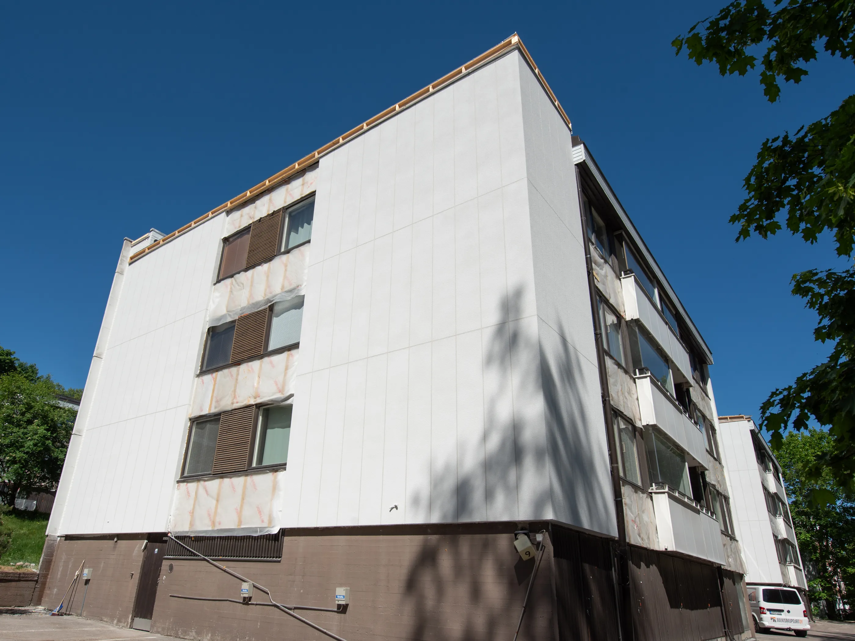 Aurinkoisena päivänä otettu alaviistokuva kerrostalosta, jonka julkisivua remontoidaan. Rakennuksen vasen ja etuosa ovat peitetty uusilla, vaaleilla pystysuorilla paneeleilla, joiden yläreunassa näkyy puurimoja. Oikealla puolella on vanhempaa julkisivua ruskeine ikkunanpuitteineen ja sälekaihtimineen, osittain peitettynä läpikuultavalla suojamateriaalilla. Rakennuksen tummanruskeassa alaosassa on putkia ja sähkökaappeja. Valkoinen pakettiauto on pysäköity oikealle. Taivas on kirkkaan sininen, ja puiden varjot lankeavat vaalealle seinälle.