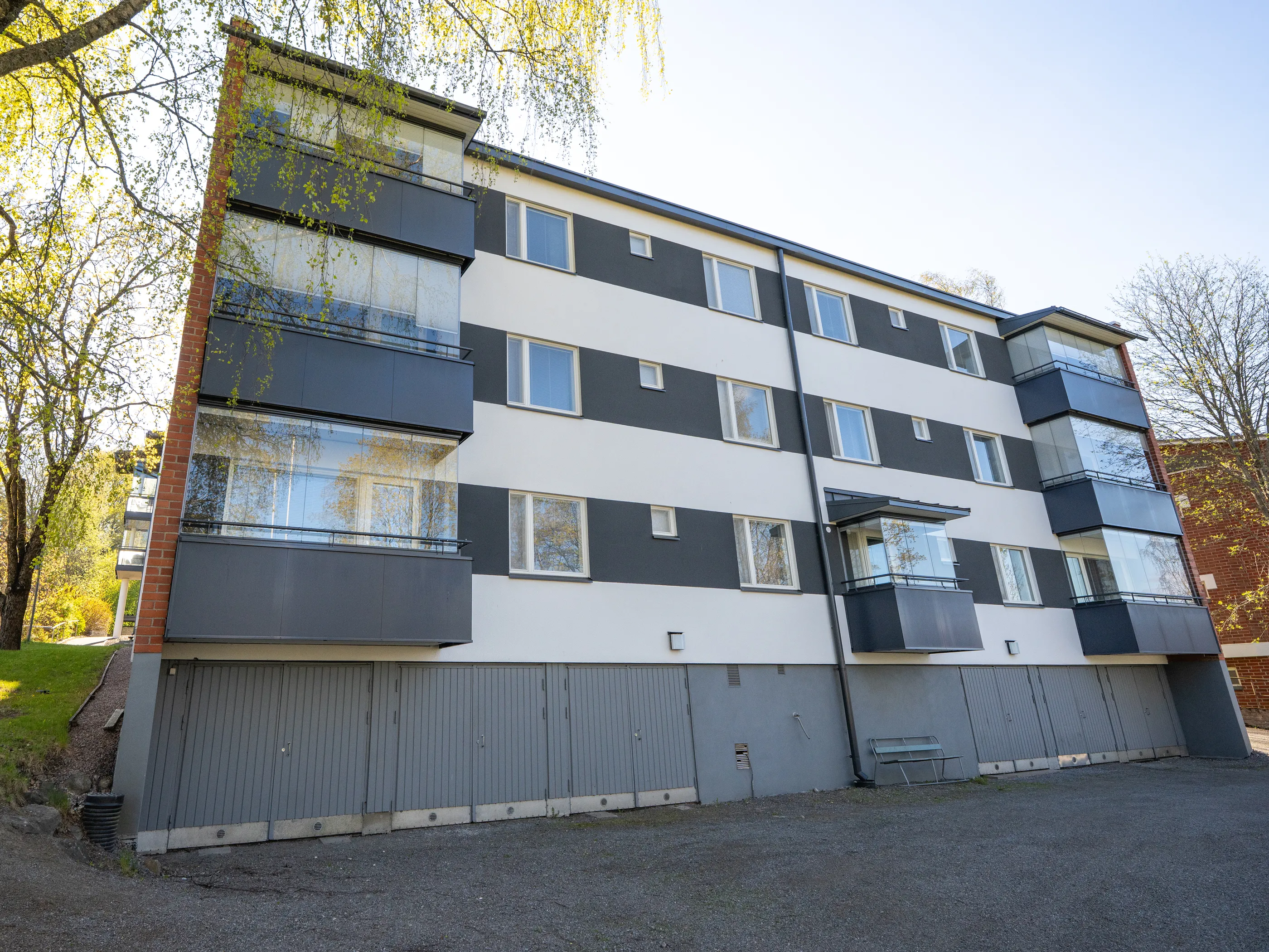 Aurinkoisena kevätpäivänä otettu kuva modernista kerrostalosta, jonka julkisivu on tummanharmaa-valkoraidallinen. Rakennuksessa on useita lasitettuja parvekkeita ja ikkunoita. Alakerroksessa on rivi harmaita ovia. Vasemmalla puolella on puita kirkkaanvihreine lehtineen, ja oikealla näkyy tiilirakennus. Etualalla on sorapintainen piha.