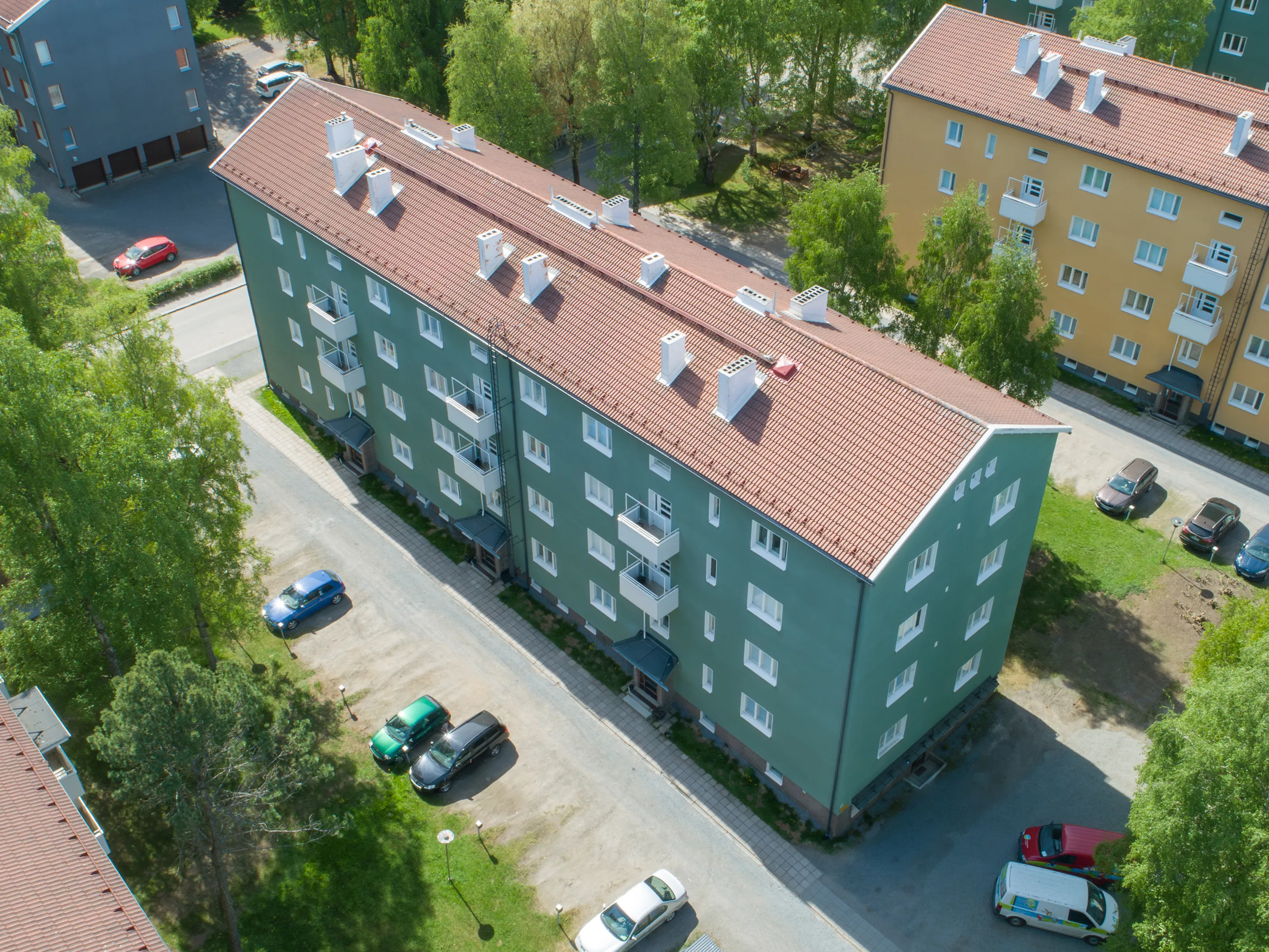 Ilmakuva pitkästä, vihreästä kerrostalosta, jossa on punaruskea tiilikatto ja valkoisia savupiippuja. Rakennuksessa on valkoiset ikkunat ja parvekkeet. Sen ympärillä on pysäköintialueita useine autoineen, sekä vihreitä puita ja nurmikkoa. Oikealla näkyy osittain keltainen kerrostalo ja vasemmalla tummanharmaa rakennus. Aurinkoinen päivä.