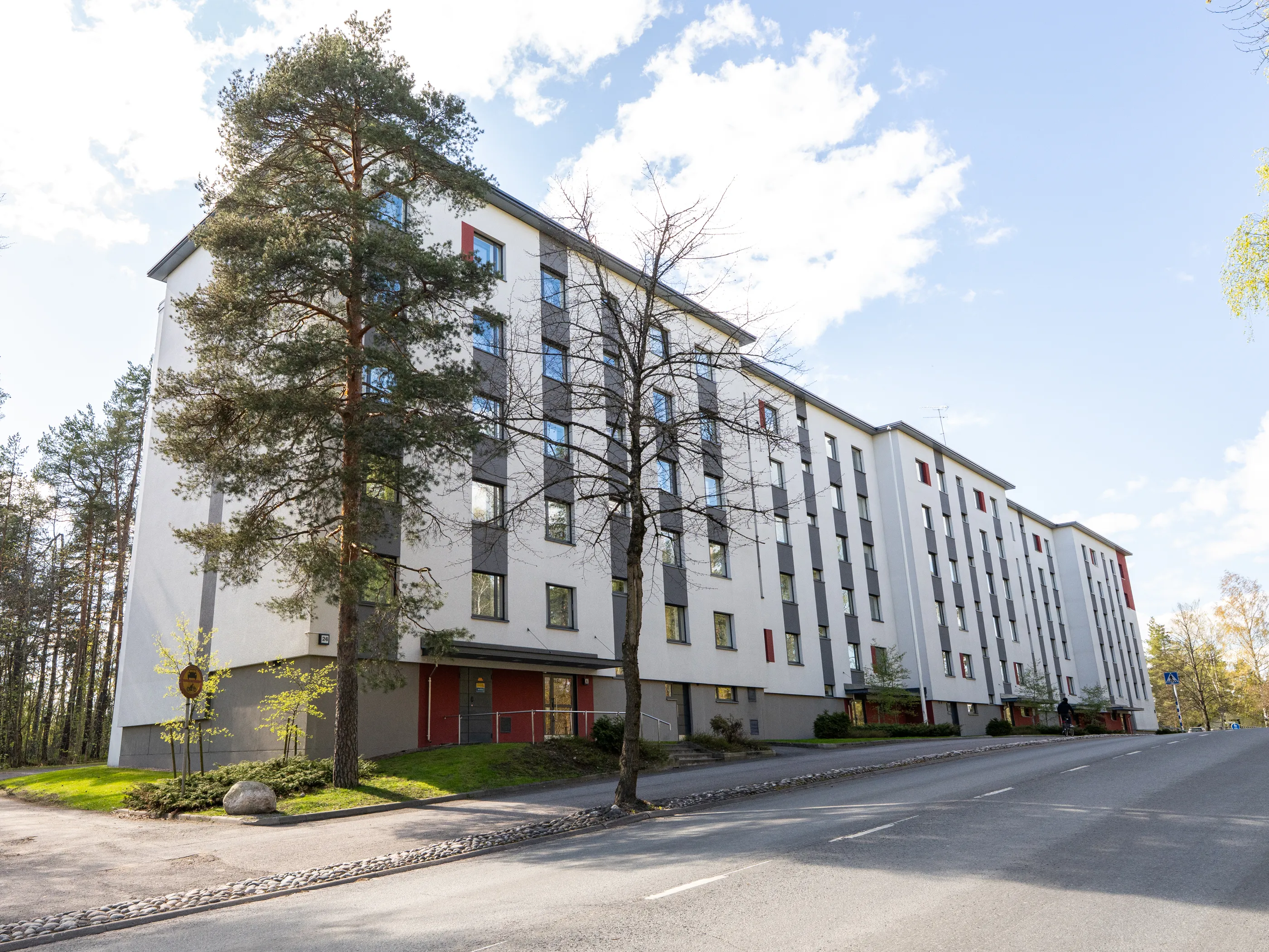 Pitkä, monikerroksinen kerrostalo, jossa on valkoiset seinät ja harmaat pystysuorat paneelit. Ikkunat heijastavat sinistä taivasta ja valkoisia pilviä. Rakennuksen edessä on mäntyjä ja muita puita, joissa on vihreitä lehtiä. Etualalla on asfalttitie kivireunuksella. Aurinkoinen päivä, ja kaukana tiellä näkyy polkupyöräilijä.