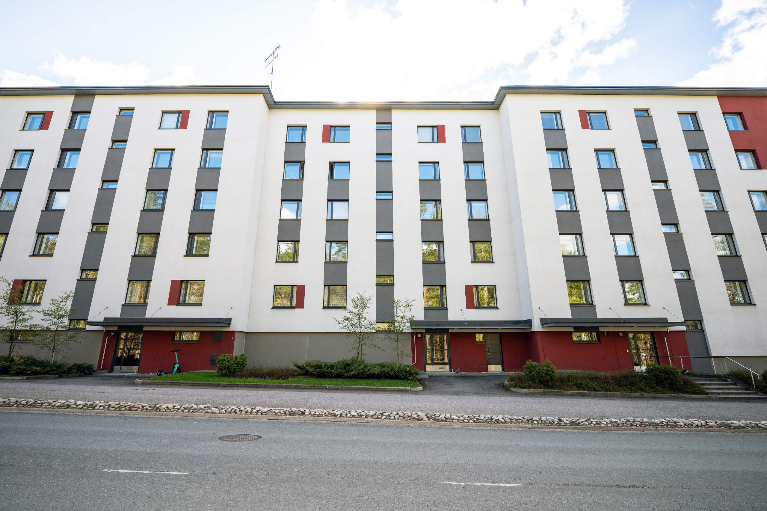 Monikerroksinen asuinrakennus, jossa on valkoisia, tummanharmaita ja punaisia julkisivuosia. Ikkunat heijastavat sinistä taivasta ja puita. Rakennuksen edessä on asfalttitie, kivireunus ja viheralue pensaineen. Oikealla näkyy turkoosi skootteri.