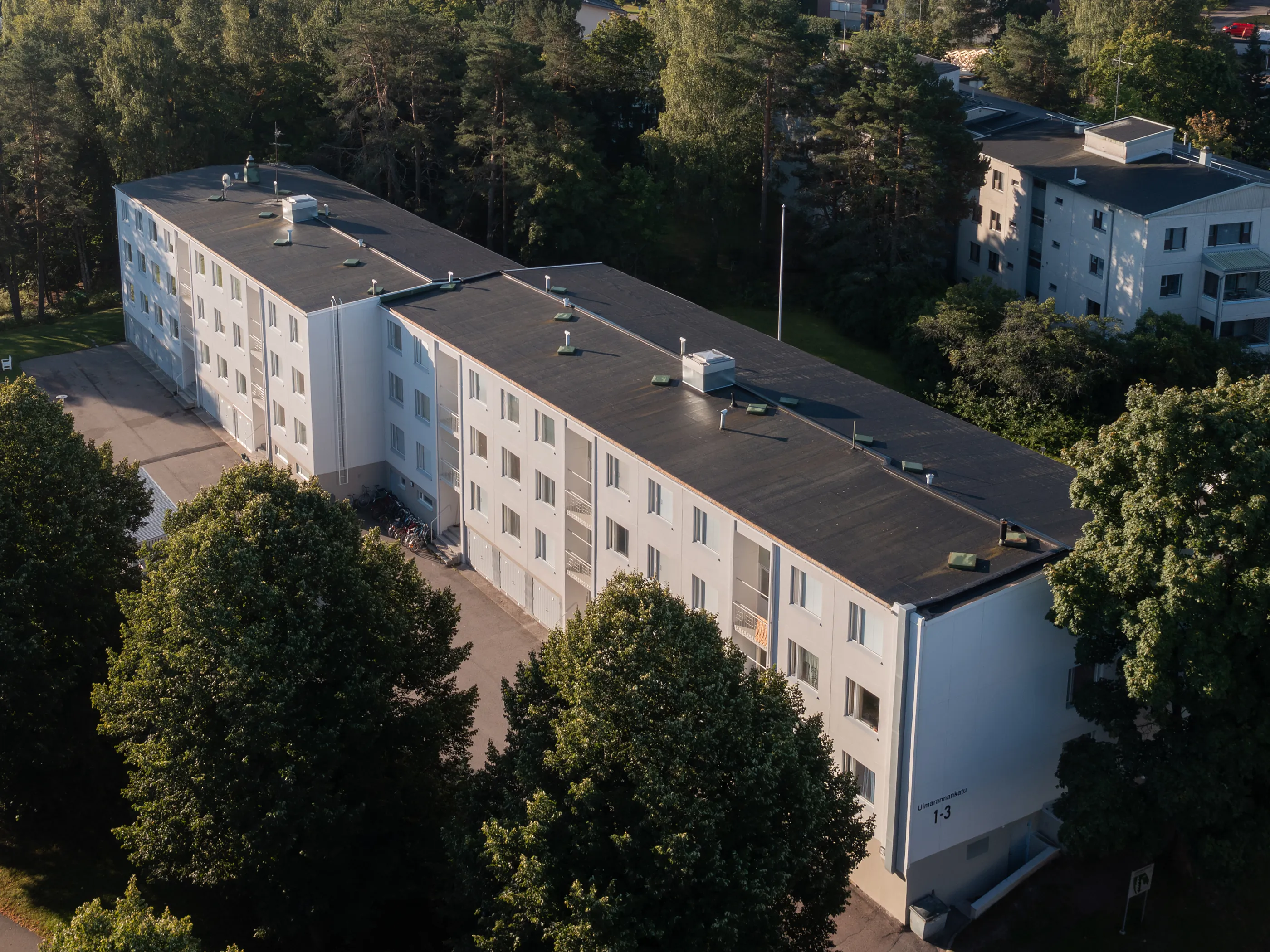 Ilmakuva pitkästä, valkoisesta kerrostalosta, jolla on tumma tasakatto. Rakennuksen ympärillä on tiheää vihreää metsää ja asfaltoitu piha, jossa on polkupyöriä. Katolla näkyy ilmastointilaitteita ja antenneja. Rakennuksen päädyssä lukee "Uimarannankatu 1-3". Aurinkoinen päivä valaisee maisemaa.