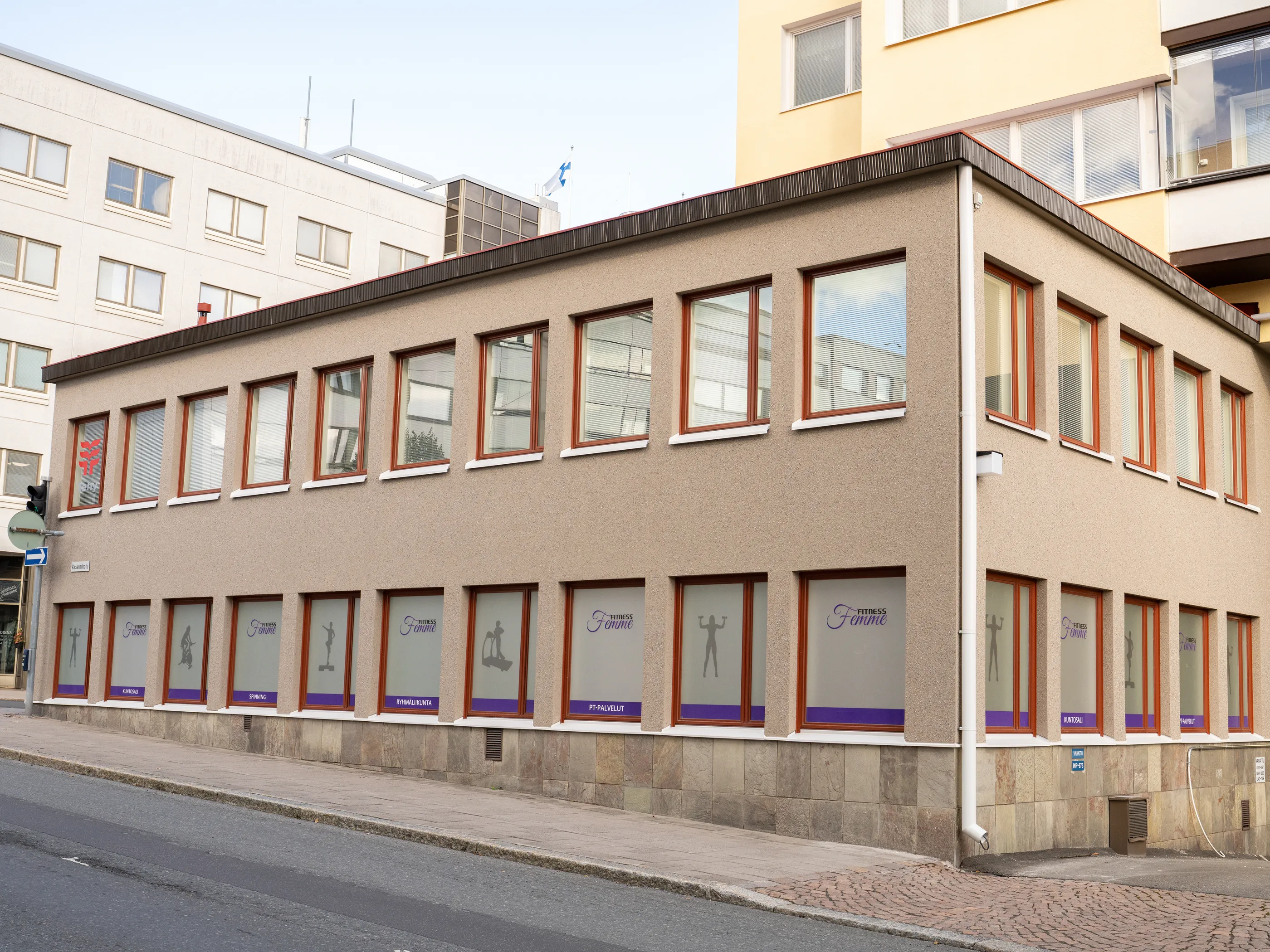 Vaaleanruskea kaksikerroksinen rakennus kadun varrella. Yläkerrassa on rivi ikkunoita ruskeilla karmeilla. Alakerran ikkunoissa on violetti "Fitness Femme" -logo, harmaita kuntoilevien ihmisten siluetteja ja palveluiden nimiä kuten "KUNTOSALI" ja "SPINNING". Rakennuksen edessä on jalkakäytävä ja asfalttitie. Taustalla näkyy muita rakennuksia, ja valkoisen rakennuksen katolla liehuu Suomen lippu.