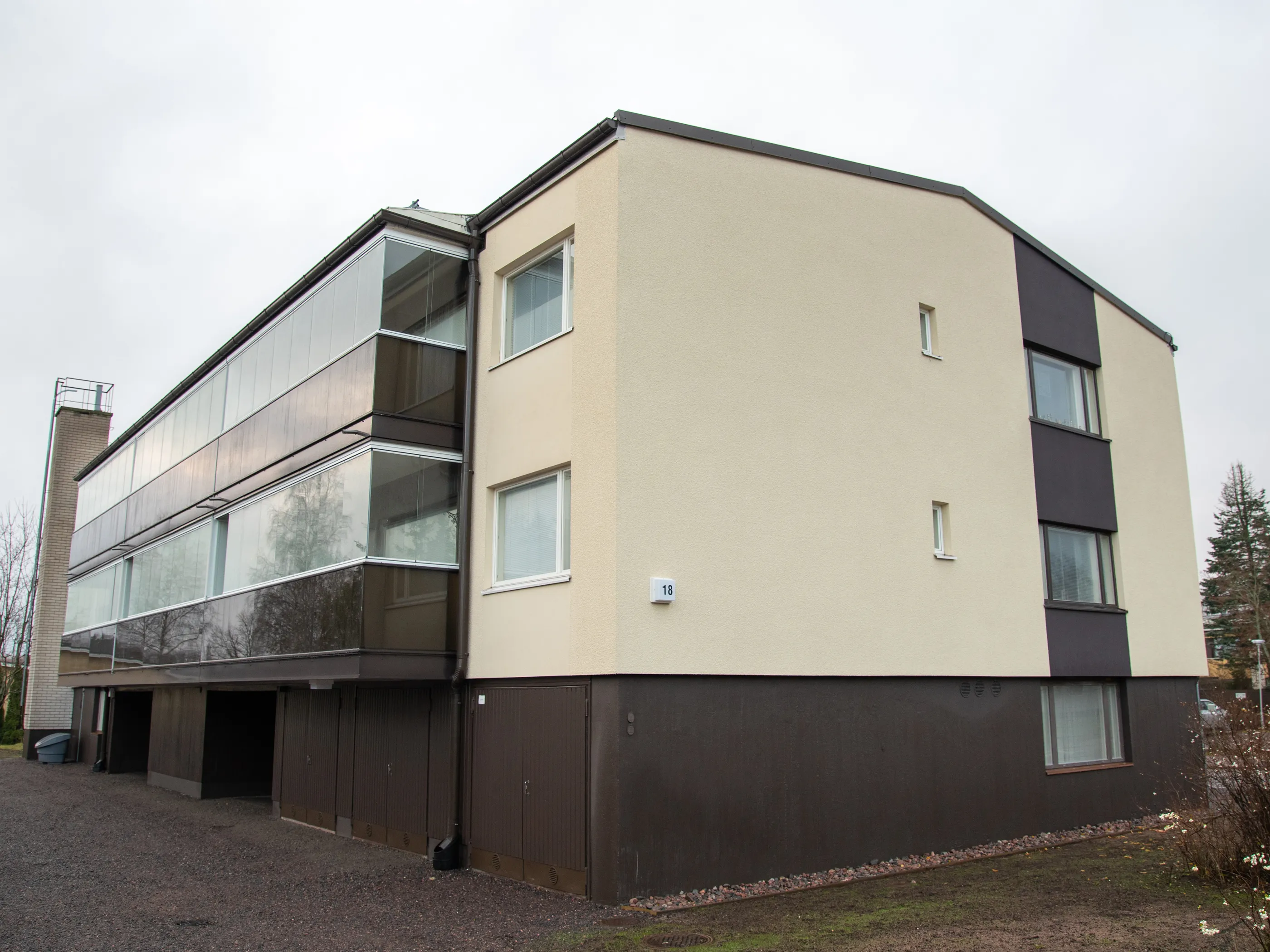 Moderni, kolmikerroksinen asuinrakennus pilvisenä päivänä. Rakennuksen yläosa on vaalean beigen värinen, ja vasemmalla puolella on suuria lasitettuja parvekkeita. Oikealla puolella on tummanruskea pystysuora paneeli ikkunoineen. Rakennuksen alaosa ja autotallin ovet ovat tummanruskeat. Vaalealla seinällä on talon numero 18. Maassa on soraa ja ruohoa, ja taustalla näkyy puita. Valo on pehmeä ja tasainen.