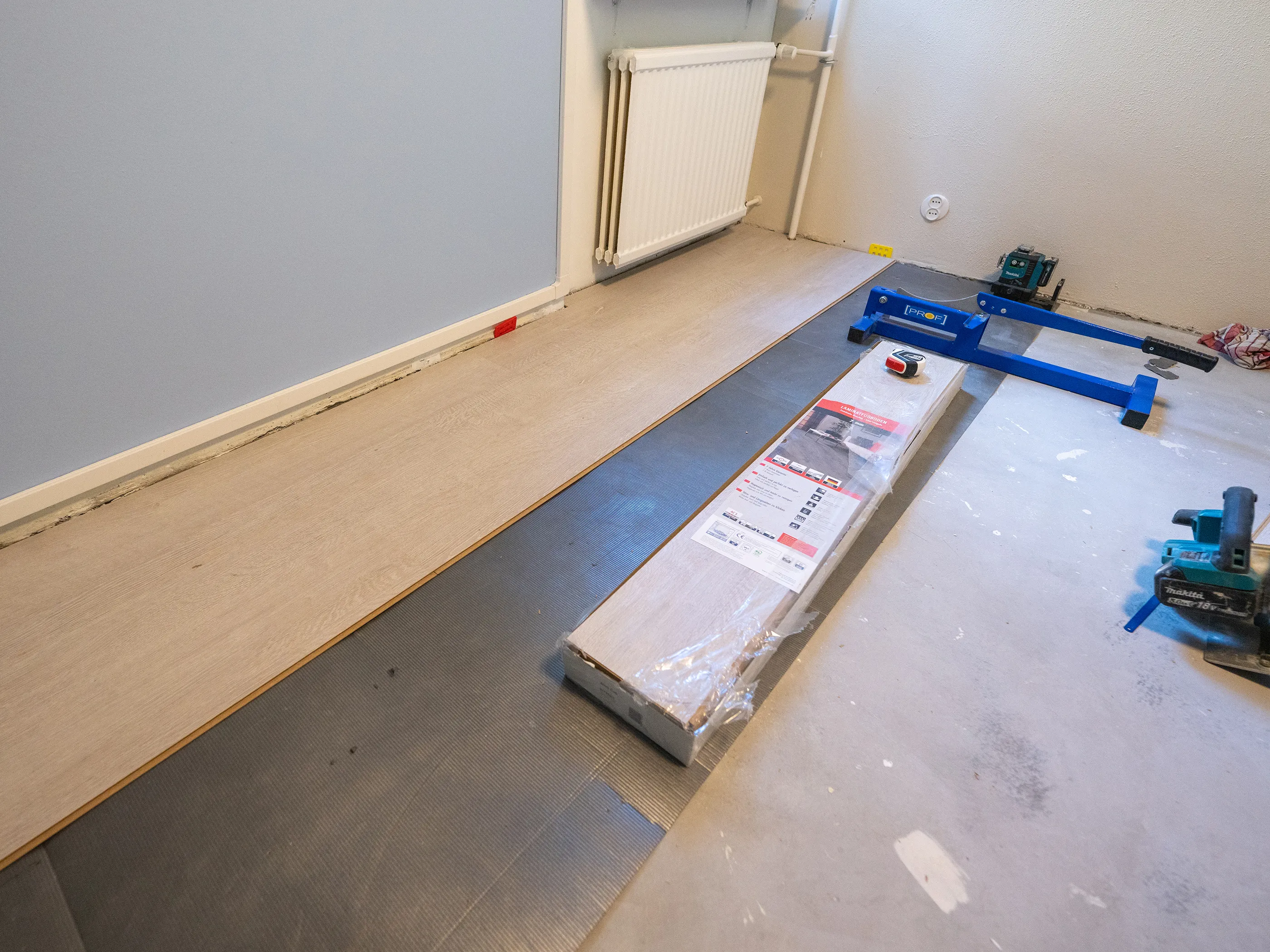 Huoneessa asennetaan vaaleaa laminaattilattiaa tummanharmaan alusmateriaalin päälle. Lattialla on paketti laminaattilankkuja, sininen asennustyökalu, vihreä laserlinjain ja mittanauha. Vasen seinä on vaaleansininen, oikea seinä on vaalea ja siinä on valkoinen patteri sekä pistorasia.