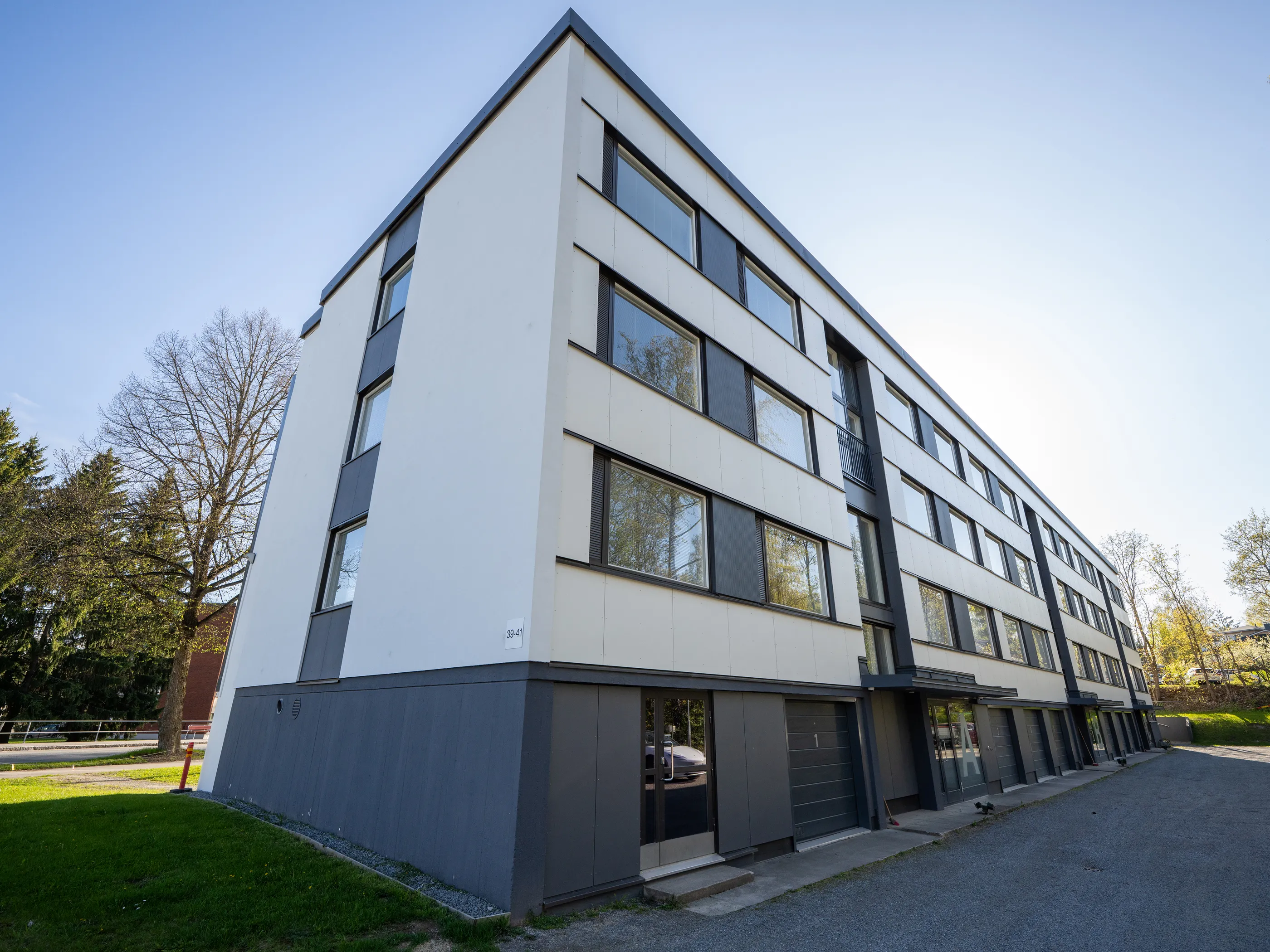 Moderni, valkoinen ja tummanharmaa kerrostalo aurinkoisessa säässä. Rakennuksen vasemmassa etukulmassa on numerointi 39-41. Useat ikkunat heijastavat ympäröiviä puita ja kirkasta sinistä taivasta. Edustalla on vihreää nurmikkoa ja tummaa sorapintaa. Taustalla näkyy puita.