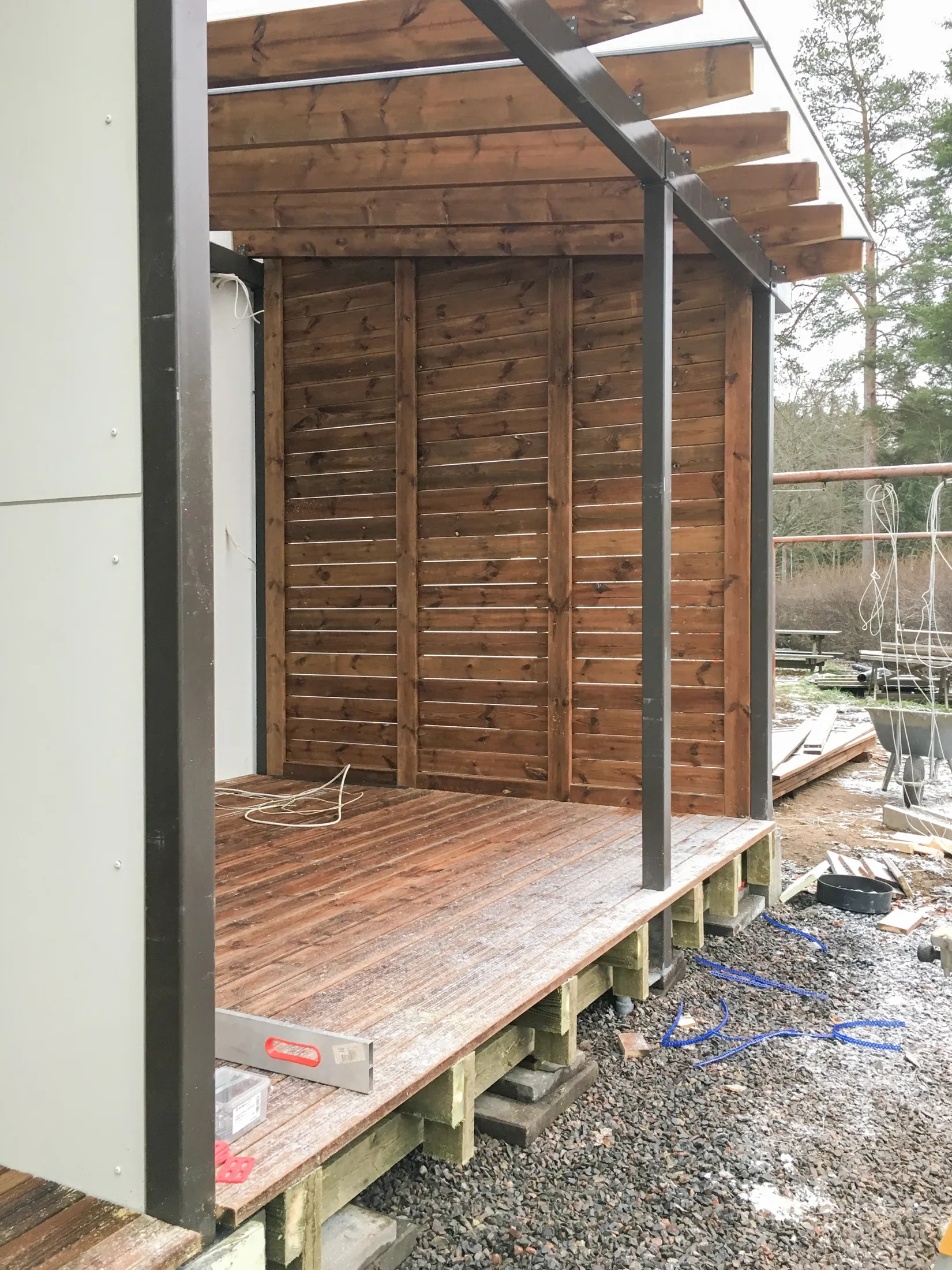 Kuva esittää rakenteilla olevaa puista terassia tai katettua oleskelutilaa. Punertavanruskea puulattia ja vaakasuorista laudoista koostuvat seinät ovat osittain kuuran peitossa. Tummanharmaat metallipylväät ja -palkit tukevat puupalkeista ja läpikuultavasta levystä muodostuvaa kattoa. Lattialla on valkoinen sähköjohto ja vatupassi. Maassa on soraa, lunta ja rakennustarvikkeita. Taustalla näkyy puita.