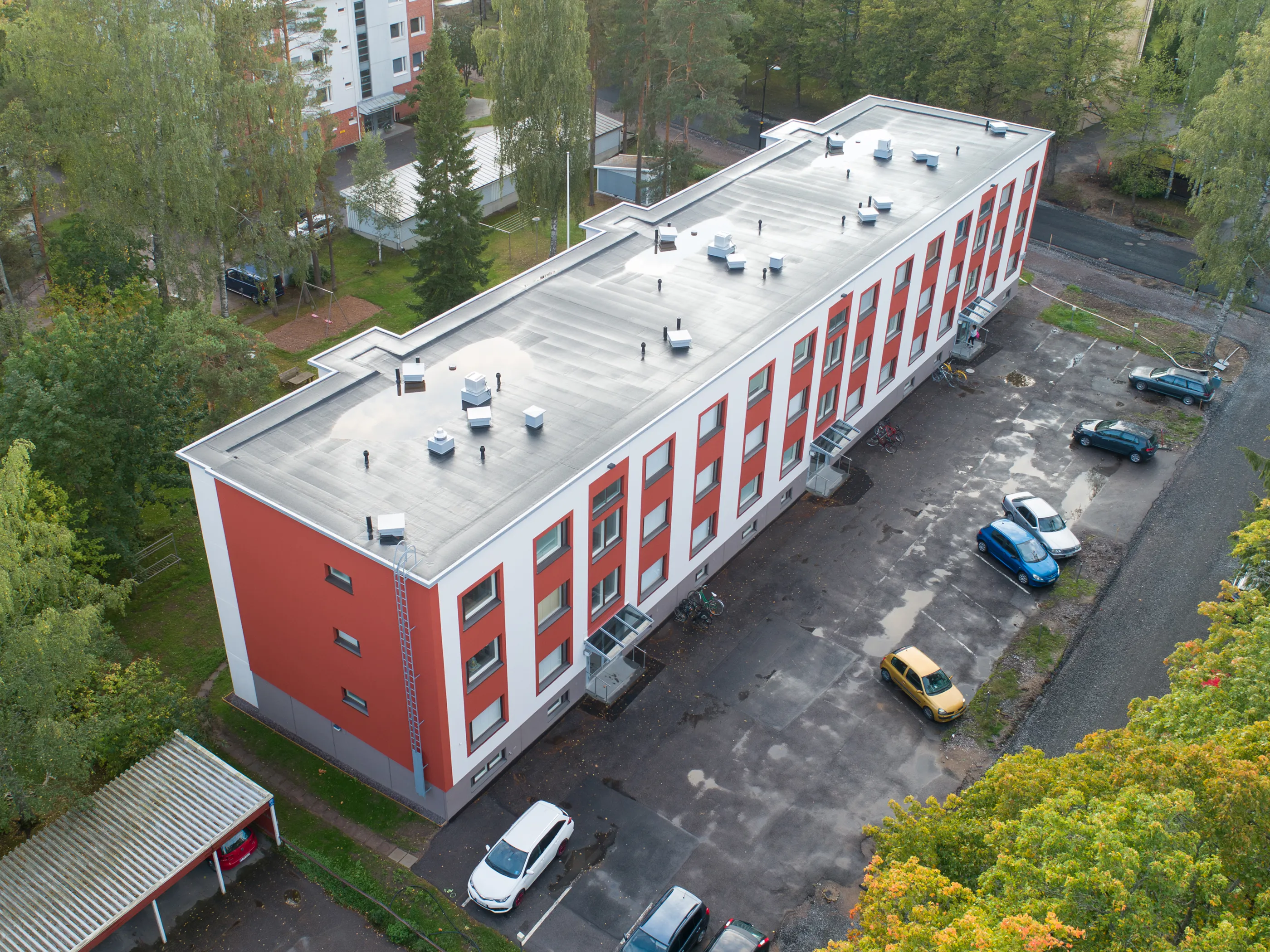 Ilmakuva pitkästä, punavalkoisesta rakennuksesta, jonka tasakatolla on ilmanvaihtolaitteita ja vesilätäköitä. Rakennuksen ympärillä on pysäköintialue useine autoineen (sininen, hopea, tummanharmaa, keltainen, valkoinen) ja vihreitä, keltaisia sekä oransseja puita. Kuvassa näkyy myös autokatos punaisen auton kanssa ja pienempiä rakennuksia. Valo on pehmeä, pilvisen päivän valo.