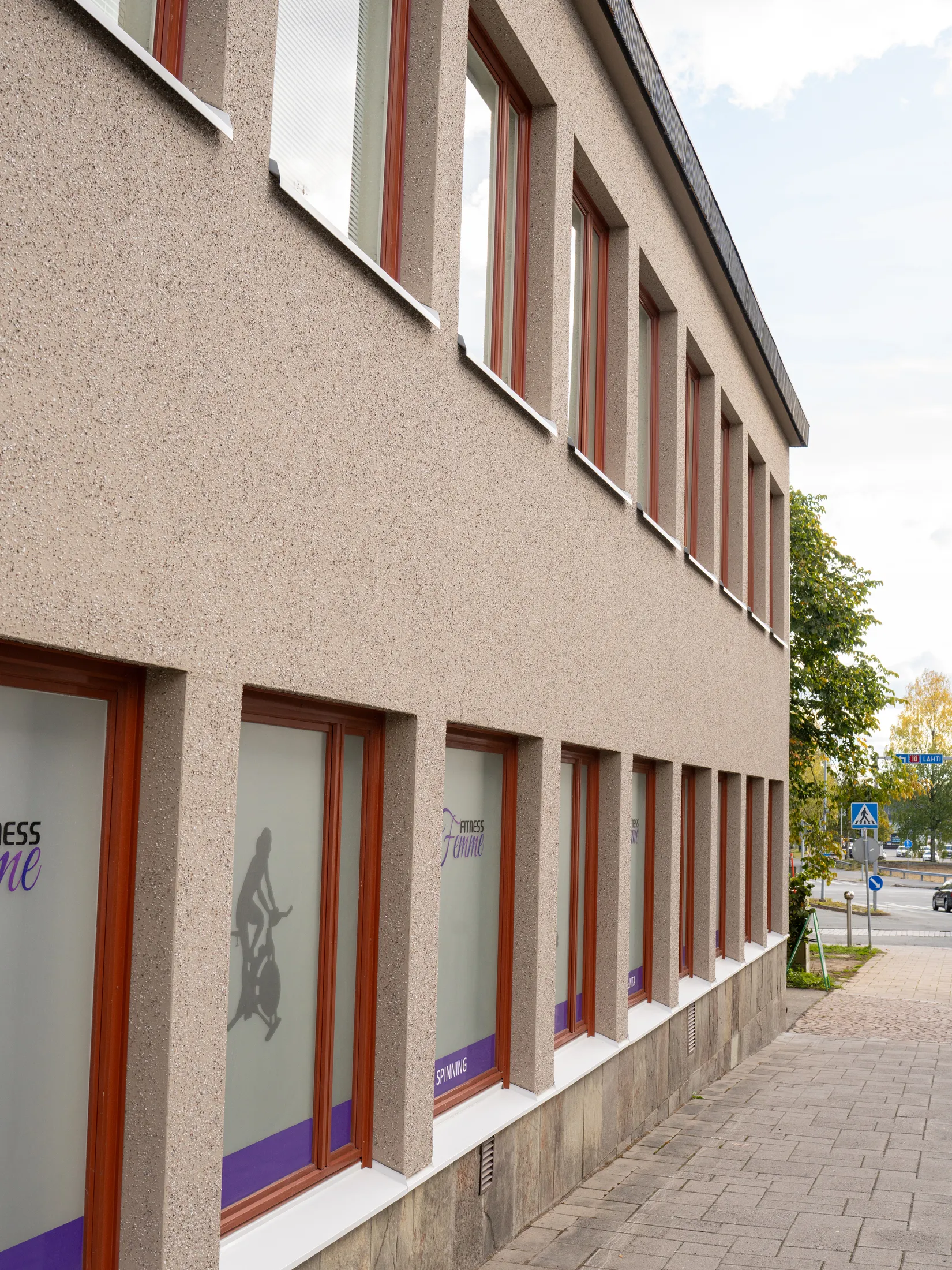 Kuva esittää rakennuksen julkisivua kirkkaassa päivänvalossa. Vaaleanruskea, rouheapintainen seinä on täynnä punertavanruskeakehyksisiä ikkunoita kahdessa kerroksessa. Alarivin ikkunoissa on himmeät lasit, joissa näkyy "FITNESS Femme" -logo, spinning-pyöräilijän siluetti ja sana "SPINNING" violetilla pohjalla. Ikkunoiden alla on kiviverhous ja valkoinen ikkunalauta. Edessä on harmaa jalkakäytävä, ja taustalla näkyy puita, tie, auto ja liikennemerkkejä, joista yksi osoittaa "LAHTI".