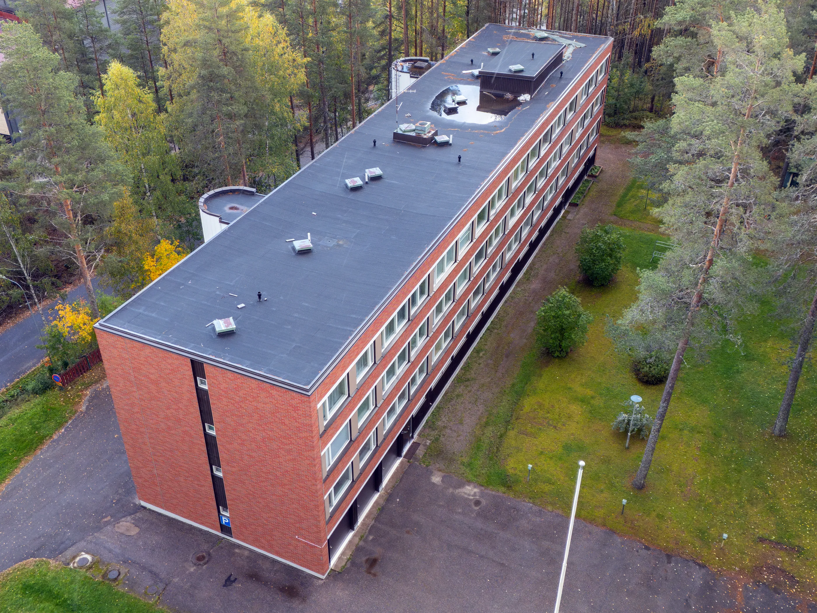 Ylhäältä kuvattu pitkä, punatiilinen rakennus, jossa on tummanharmaa tasakatto. Katolla näkyy ilmanvaihtoputkia, laitteita ja suuri vesilätäkkö. Rakennuksen pitkällä sivulla on rivi ikkunoita ja päädyssä sininen P-kyltti. Rakennusta ympäröi vihreä nurmikko, asfalttipolkuja, lipputanko, katuvaloja ja runsaasti puita, mukaan lukien mäntyjä ja keltaisia syksyisiä lehtipuita. Valo on pehmeä ja tasainen.