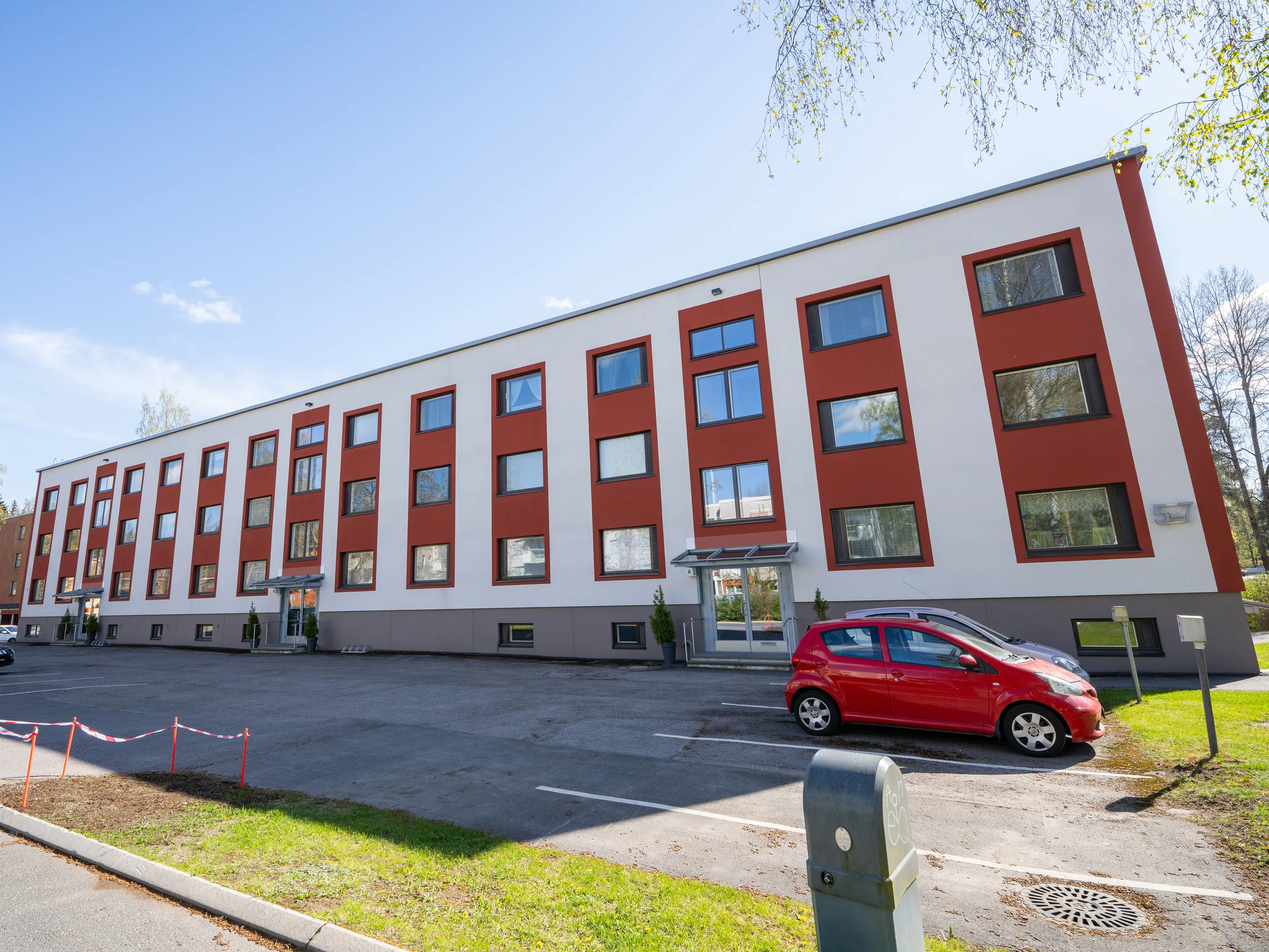 Pitkä, valko- ja punaruskearaidallinen rakennus aurinkoisessa säässä. Rakennuksessa on useita ikkunoita, jotka heijastavat taivasta ja puita. Edessä on asfalttiparkkipaikka, jossa on punainen auto ja latauspiste. Taustalla näkyy sininen taivas ja vihreää kasvillisuutta.
