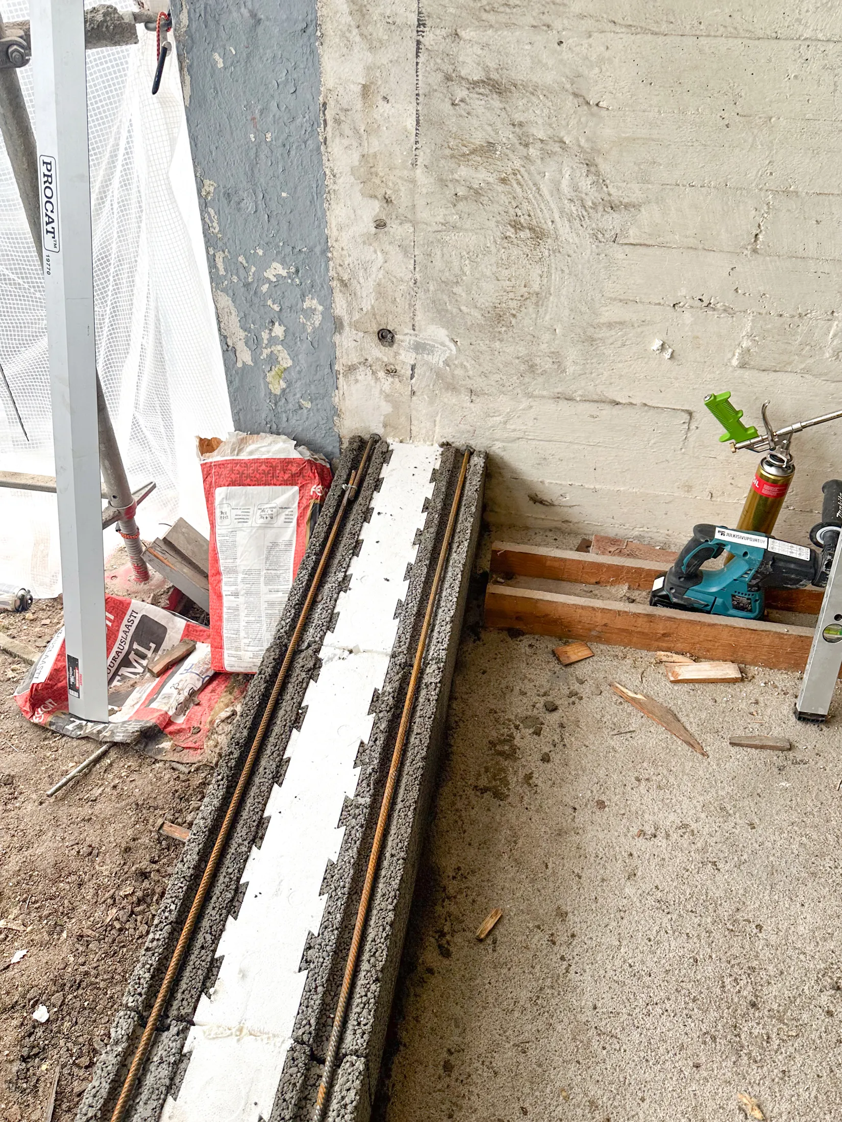 Kuva rakennustyömaalta, jossa näkyy betonia, rakennustarvikkeita ja työkaluja. Vasemmalla on valkoinen rakennustelineen pylväs ja valkoista verkkoa. Maassa on punavalkoisia laastipusseja. Keskellä on pitkä betonipalkki, jossa on harjateräkset ja valkoinen sahalaitainen eriste. Oikealla on vaalea betoniseinä, puulankkuja, turkoosi Makita-pistosaha ja vihreällä annostelupistoolilla varustettu rakennusvaahtopullo.