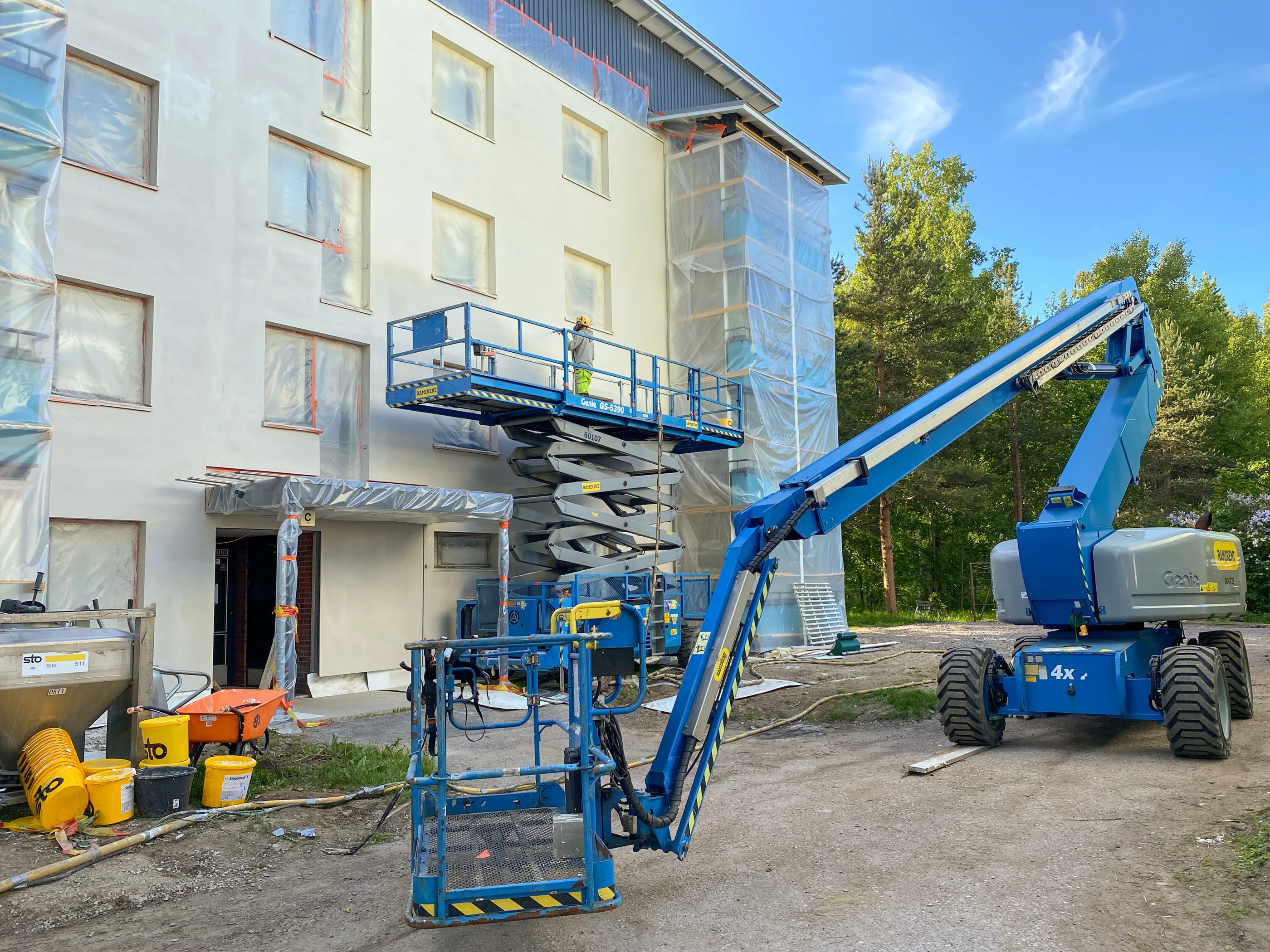 Vaalea rakennus on remontissa, sen ikkunat on peitetty muovilla. Sininen saksinostin on nostettu ylös, ja sen korissa on työntekijä keltaisessa kypärässä ja huomiovaatteissa. Oikealla seisoo toinen sininen puominostin. Maassa on rakennustarvikkeita, kuten siilo, keltaisia ämpäreitä ja oranssit kottikärryt. Taustalla on vihreitä puita ja kirkas sininen taivas.