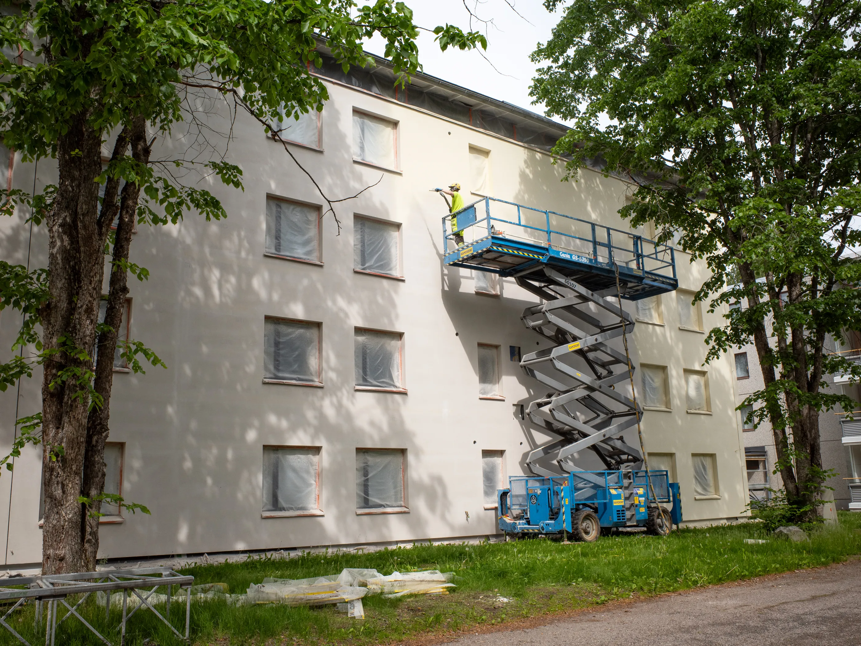 Aurinkoisena päivänä työntekijä maalaa vaaleaa rakennuksen julkisivua sinisellä saksinostimella. Rakennuksen ikkunat on peitetty suojamuovilla. Vihreät puut reunustavat rakennusta ja heittävät varjoja seinälle. Etualalla on ruohoa ja pakattuja rakennustarvikkeita.