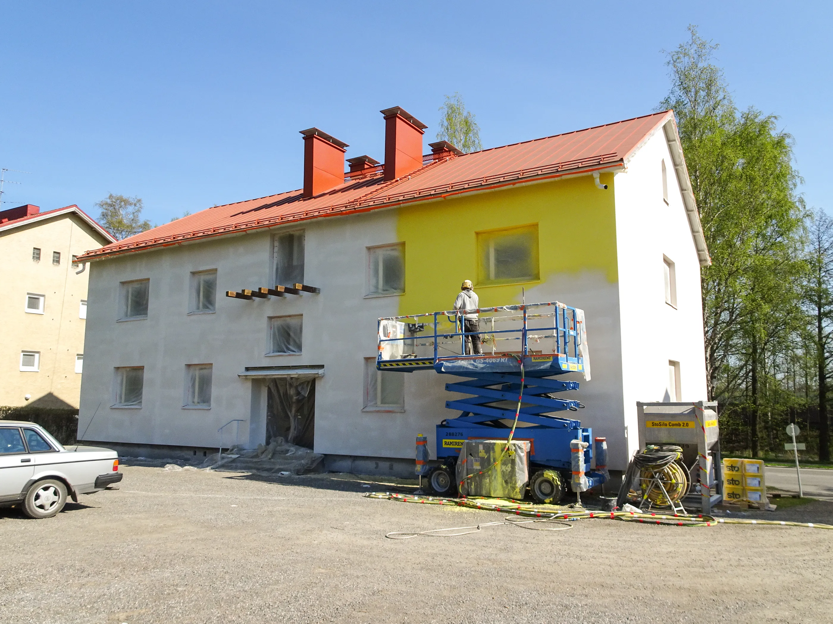 Aurinkoisena päivänä kaksikerroksista rakennusta maalataan ulkoa. Rakennuksen vasen puoli on vaaleanharmaassa pohjamaalissa, ja oikea puoli on jo maalattu kirkkaan keltaiseksi. Maalari seisoo sinisellä saksinostimella ja ruiskuttaa keltaista maalia seinään. Rakennuksessa on punainen peltikatto ja punaisia savupiippuja. Ikkunat on suojattu muovilla. Maassa on soraa, ja siinä on maalaustyömaalle kuuluvia letkuja, siilo ja muita tarvikkeita. Taustalla näkyy toinen rakennus, vihreitä puita ja kirkas sininen taivas. Kuvan vasemmassa alareunassa on hopeinen henkilöauto.