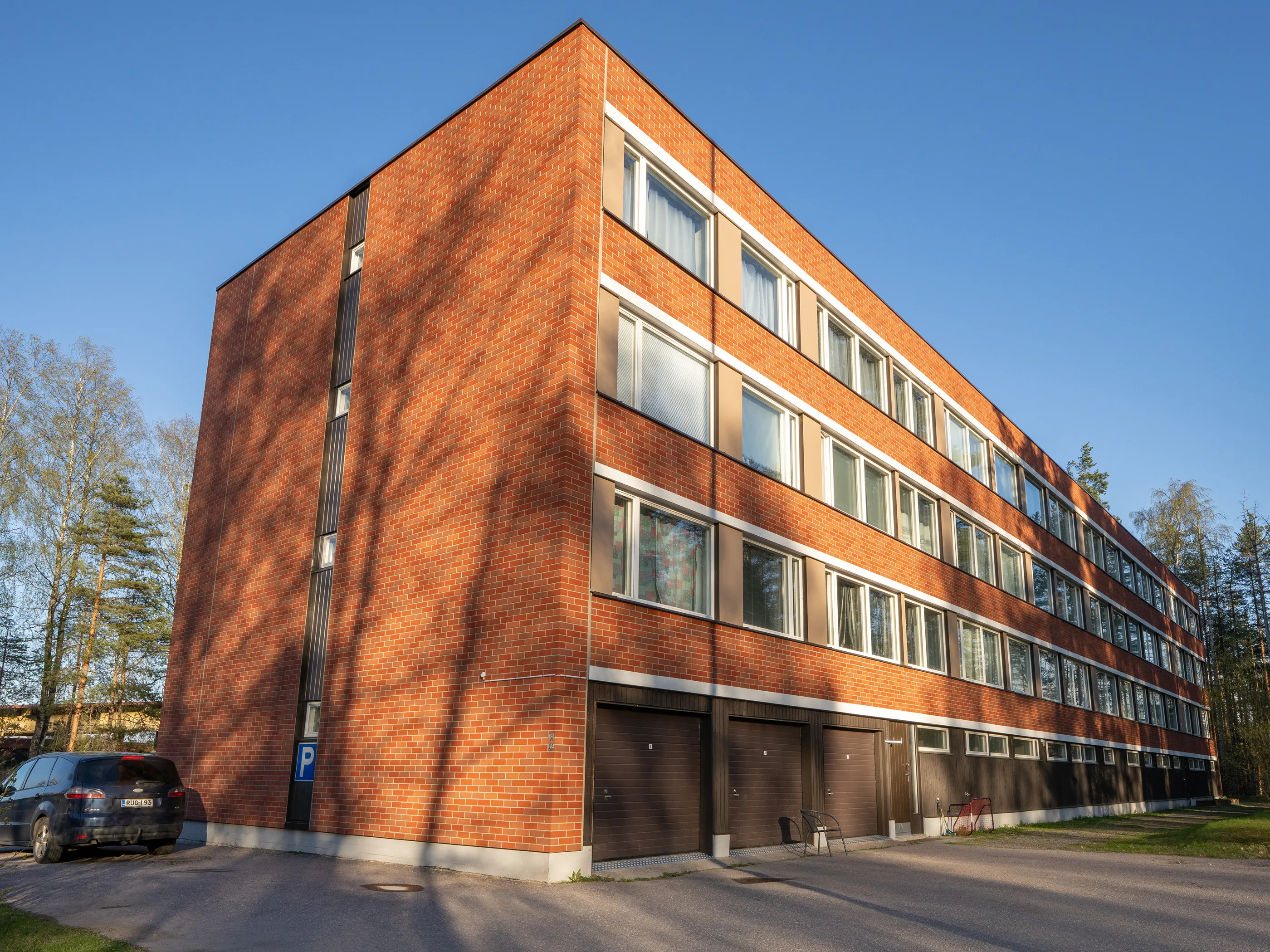 Aurinkoinen kuva punatiilisestä kerrostalosta, jossa on useita valkoisia ikkunoita ja tummanruskeita autotallin ovia alakerroksessa. Rakennuksen vasemmalla puolella on pysäköity tummansininen auto ja P-kyltti. Puista lankeaa pitkiä varjoja rakennuksen julkisivulle ja asfalttipihalle. Taivas on kirkkaan sininen ja puita näkyy rakennuksen ympärillä.