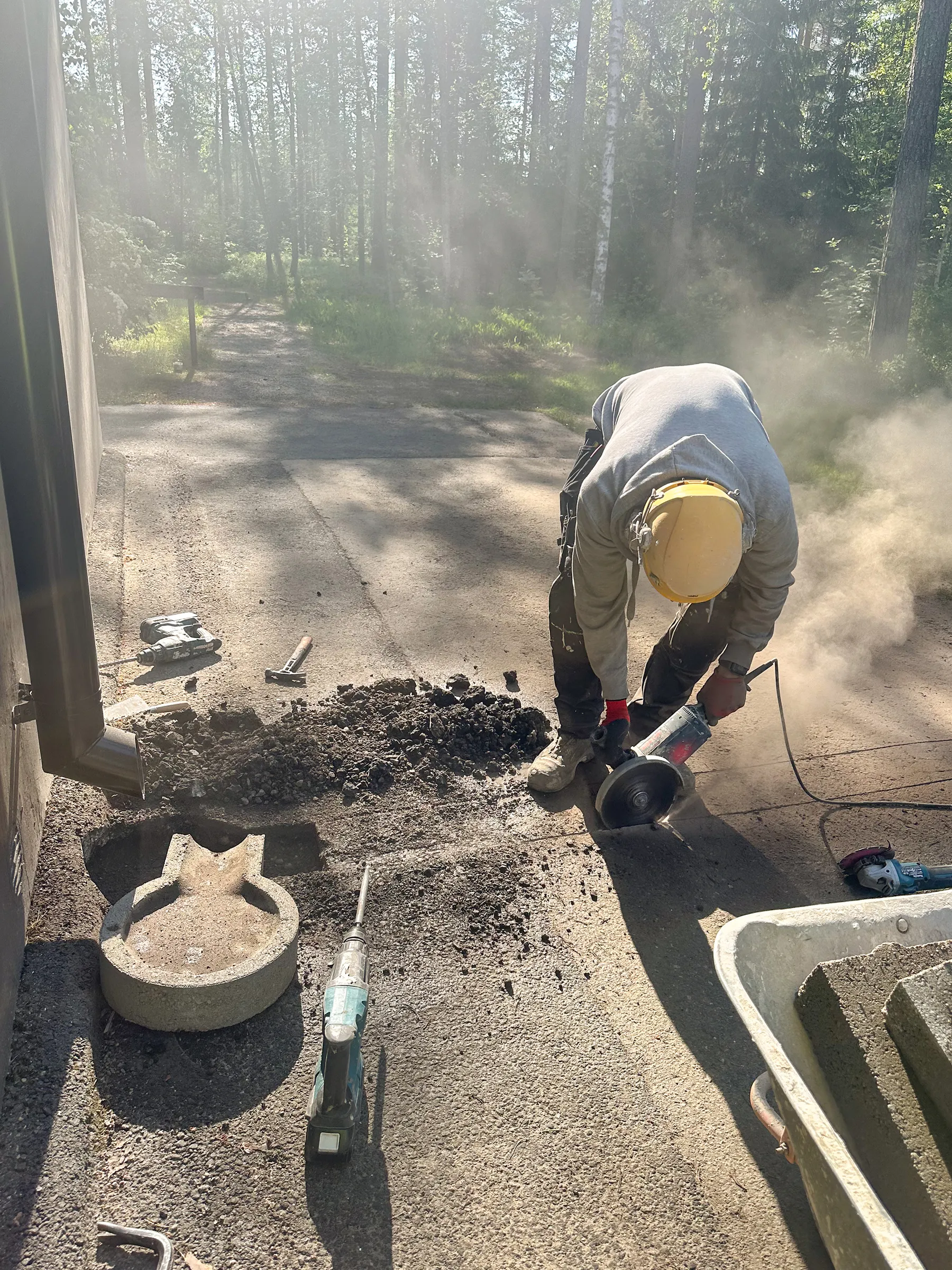 Mies keltaisessa kypärässä ja harmaassa hupparissa leikkaa asfalttia kulmahiomakoneella, nostattaen paljon pölyä. Maassa on työkaluja, kuten purkupiikkikone, vasara ja sadevesikourun alla oleva betoninen valuma-allas. Oikealla näkyy kottikärryt betoniharkkoineen. Taustalla on aurinkoinen metsäpolku ja puita.