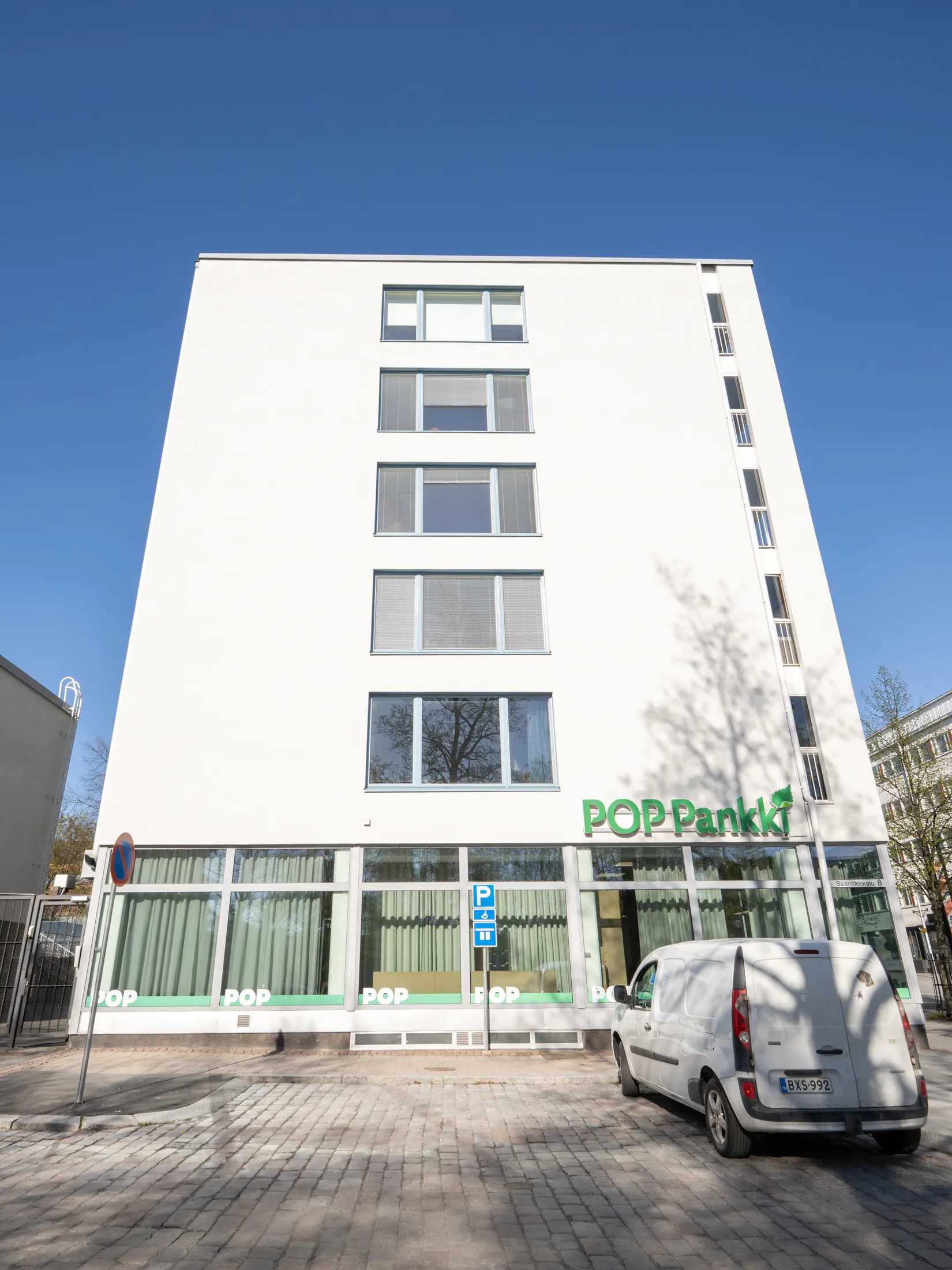 Korkea, valkoinen rakennus kirkkaan sinisen taivaan alla. Rakennuksen alaosassa on vihreä 'POP Pankki' -kyltti ja suuret ikkunat, joissa heijastuu puita. Edessä on mukulakivinen piha, jossa seisoo valkoinen pakettiauto. Aurinko paistaa kirkkaasti, luoden puun varjoja rakennuksen seinälle ja maahan.