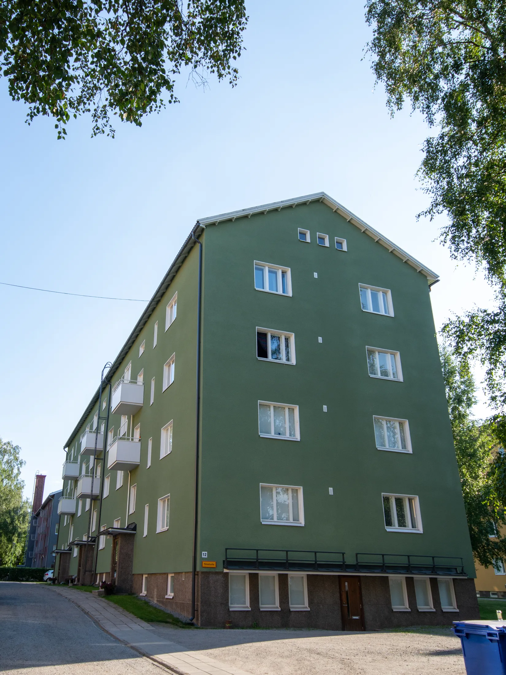 Aurinkoisena päivänä otettu kuva vihreästä kerrostalosta, jossa on valkoiset ikkunat ja parvekkeet. Rakennuksen alaosassa on tumma, kivimäinen sokkeli. Oikealla seinässä on keltainen kyltti numerolla 12 ja tekstillä 'Pelastustie'. Kuvan yläreunassa ja oikealla puolella näkyy vihreitä puunlehtiä. Edustalla on asfalttitie ja jalkakäytävä, ja oikealla alhaalla sininen roska-astia. Taustalla vasemmalla näkyy toinen rakennus.